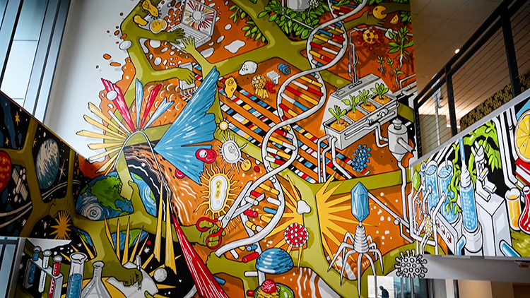 Part of a colorful wall mural depicting notable scientific contributions from the JGI. The art wraps around the mezzanine area.