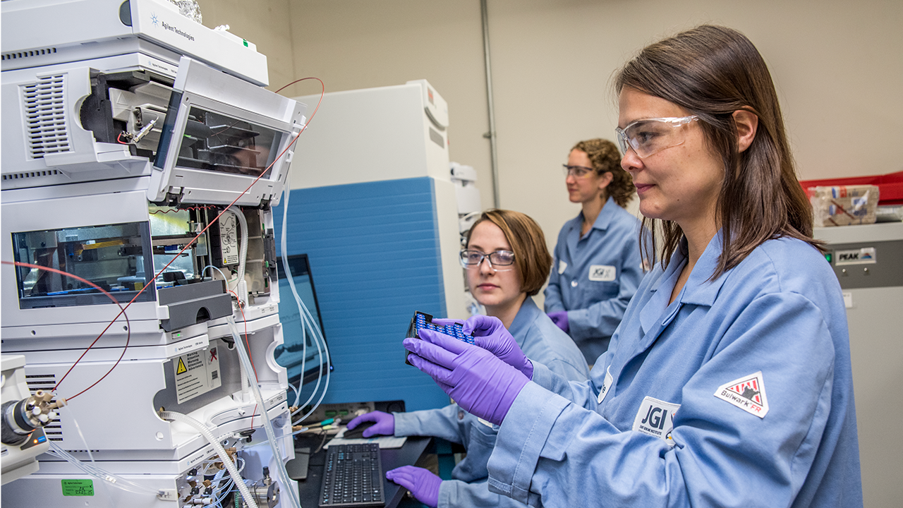 Three individuals in lab coats, safety goggles and purple gloves stand in front of scientific equipment
