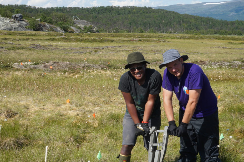 Two researchers stand in a grassy field operating grey metal coring equipment.