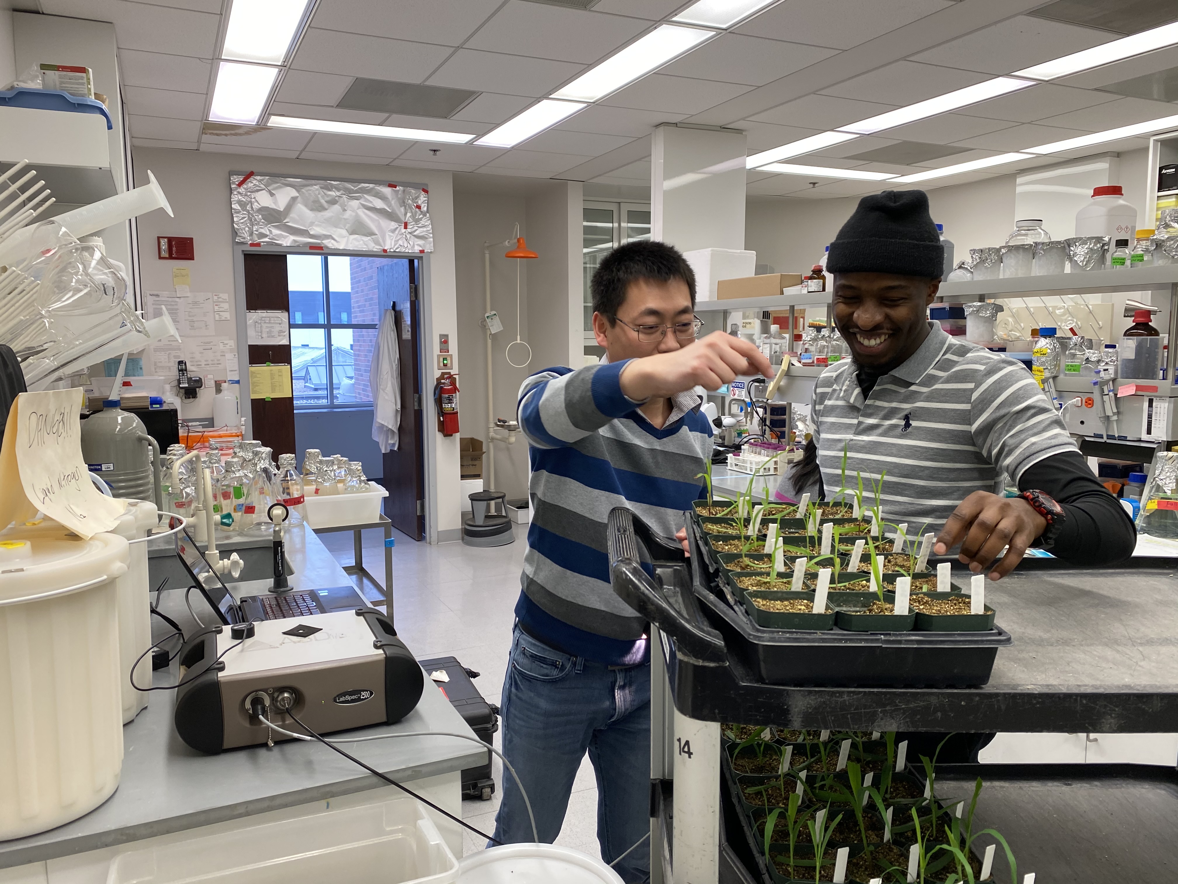 Two researchers work in a science lab with a cart of small plants.