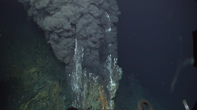 An underwater photo with a blue backdrop and a mineral formation erupting black, mineral-rich fluid.