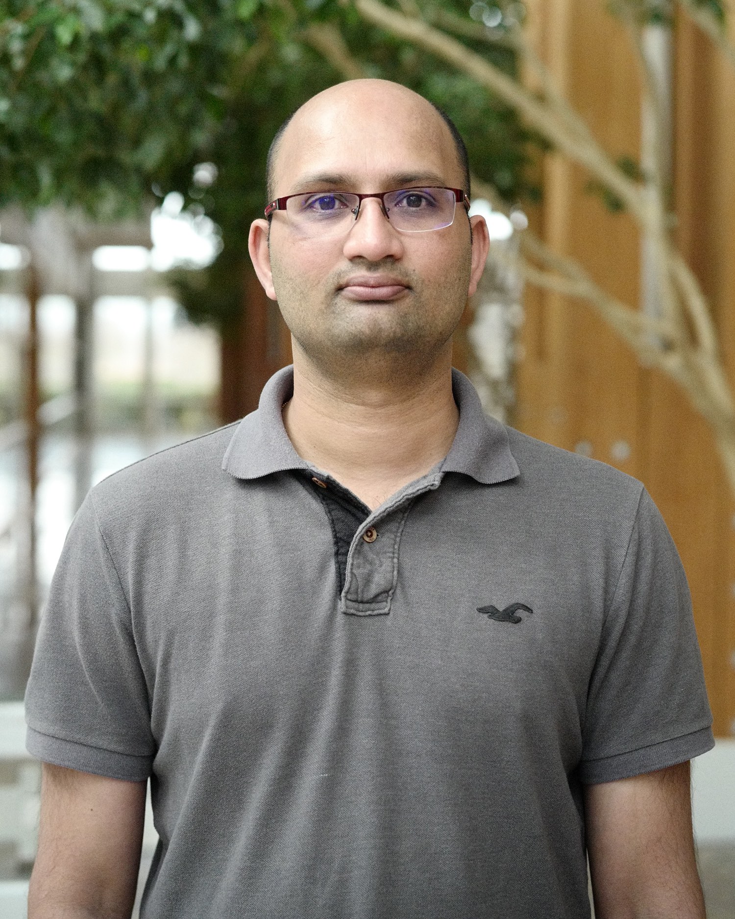 A scientist wears glasses and a grey polo in his head shot.