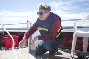 A scientist kneels on a boat to pour water samples into a large plastic jar.