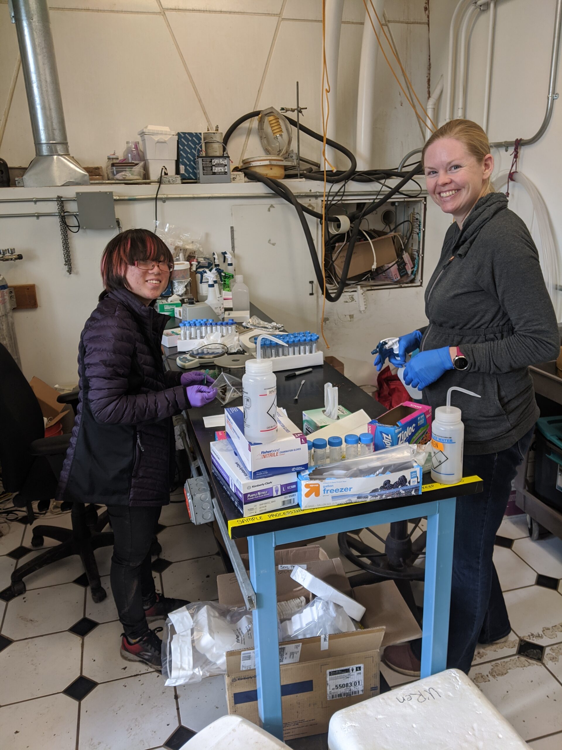 Two scientists in a laboratory at Biosphere 2, smiling as they process samples at a workbench filled with various lab equipment and supplies.