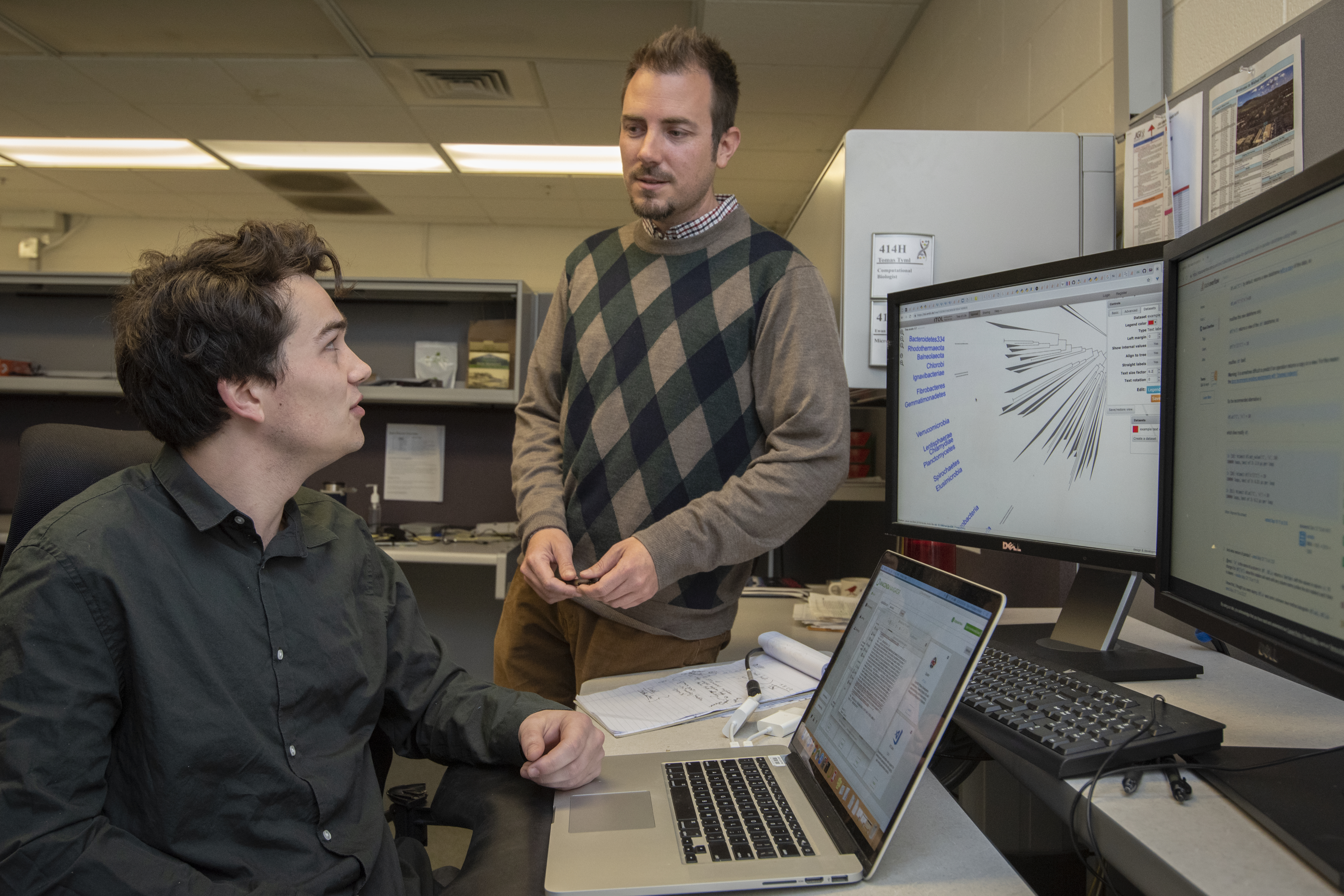 Two indviduals talk, one staneding while the other sits in front of a laptop and monitor