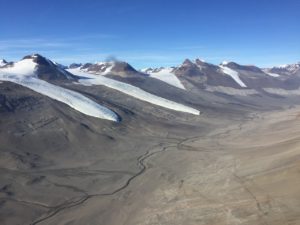 Against a blue sky are several snow-topped peaks above a dry brown valley in Antarctica.