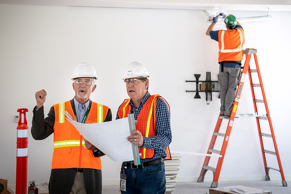 two people in construction gear talking. A person is behind them on a ladder