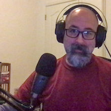 A man with a beard and glasses wears headphones in front of a microphone.