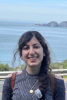 long-haired woman in cutout sweater and brown backpack smiles in front of an ocean view