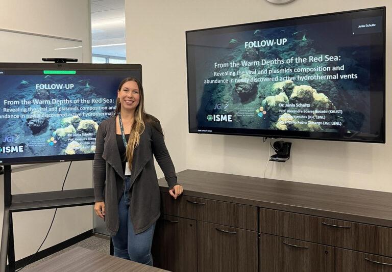 Júnia Schultz standing in a conference room in front of a TV showing her presentation