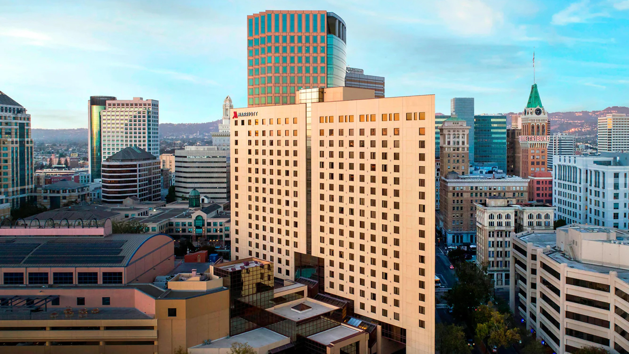 View of a city skyline featuring the Oakland Marriott hotel and a mix of modern and historic buildings, with a colorful sunset sky.