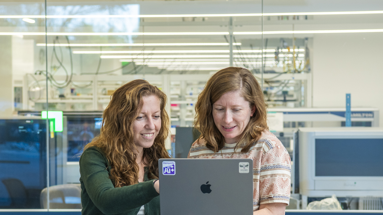 Two individuals look at a laptop in hand while the other points to the screen. A lab setting is visible in the backdrop.