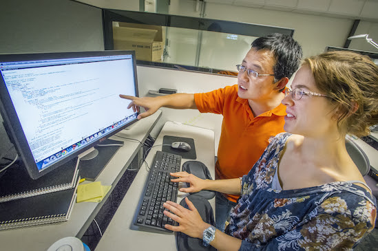 Two individuals sit at a computer. The person on the left is pointing at a computer screen offering instruction