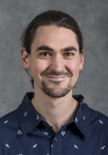 An individual with dark hair and a slight beard and mustache smiles at the camera