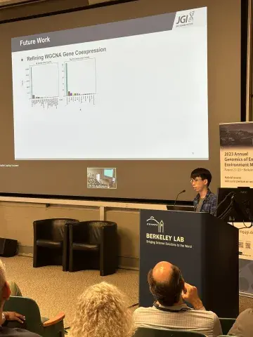 A UC Merced Intern standing at a podium presenting their work on a large screen