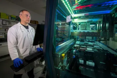 An individual in a lab coat, gloves and goggles stares into a large lit machine