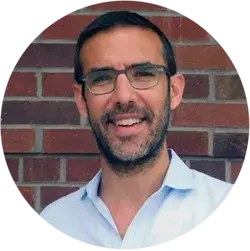 Bearded person in glasses and white shirt against a brick background.