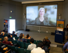 A woman presents virtually, visible from a large projector screen before her audience