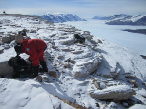 A researcher in a red snowsuit works on sampling rocks in a snowy landscape.