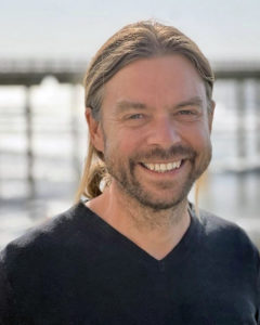 Jack Gilbert smiles in front of a blurred ocean background