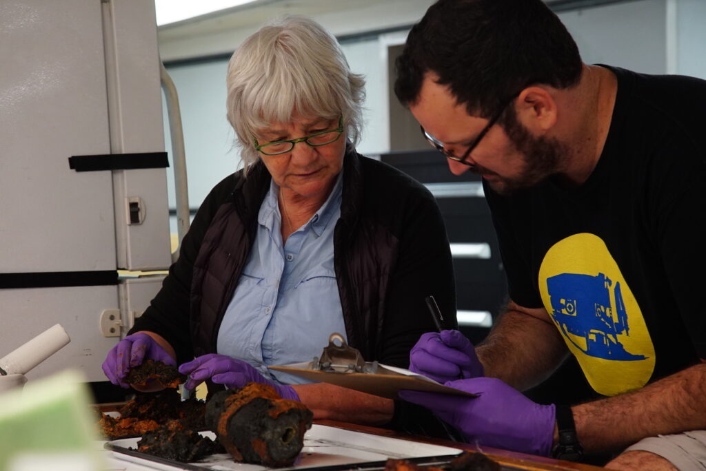 Two researchers take a look at a rock sample that is black and rust-colored.