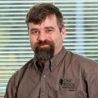 Chris Schadt with dark hair and beard and wearing a brown shirt with a logo over the left breast pocket. He smiles while standing in front of green window blinds