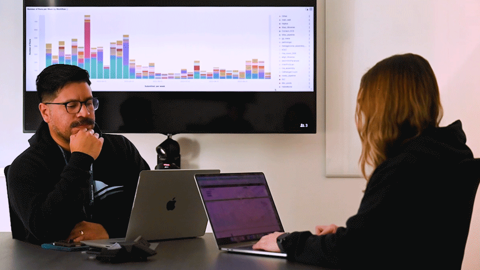 Two bioinformatics researchers sit together at a conference room table while a graph cycles through different data visualizations on the screen above them.