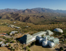 An aerial view of the Biosphere 2 facility, showing its distinct glass and steel structures set against the backdrop of rugged desert mountains and vast, arid landscape.