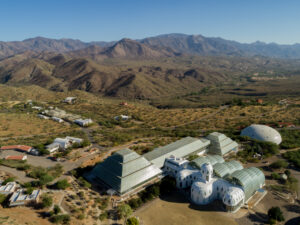 An aerial view of the Biosphere 2 facility, showing its distinct glass and steel structures set against the backdrop of rugged desert mountains and vast, arid landscape.