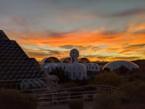 A stunning sunset over the Biosphere 2 facility, featuring its iconic geodesic domes and pyramid structures silhouetted against a vibrant, multicolored sky.