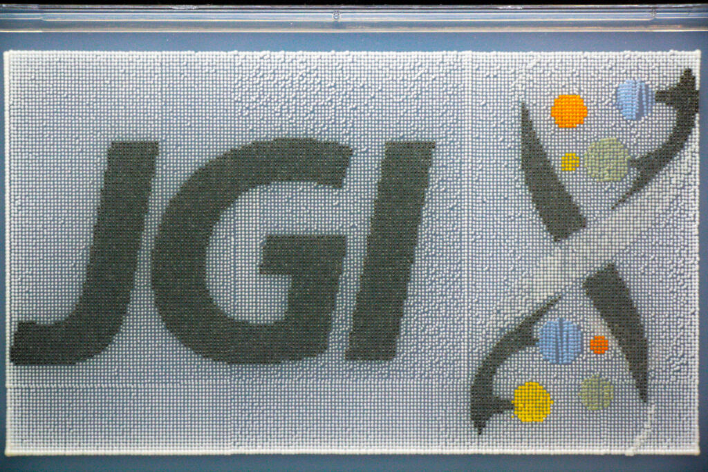 A mosaic image with the letters 'JGI' prominently displayed on the left. On the right, there is a stylized DNA double helix with several colored dots (yellow, orange, green, and blue) along its length.