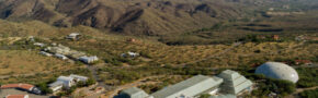 An aerial view of the Biosphere 2 facility, showing its distinct glass and steel structures set against the backdrop of rugged desert mountains and vast, arid landscape.