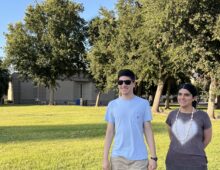 A photo of two smiling individuals standing outdoors on a grassy area in front of a building with trees in the background.