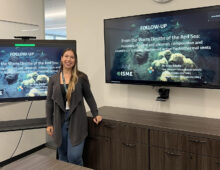 Postdoc Junia Schultz standing in a conference room with her presentation on a tv screen